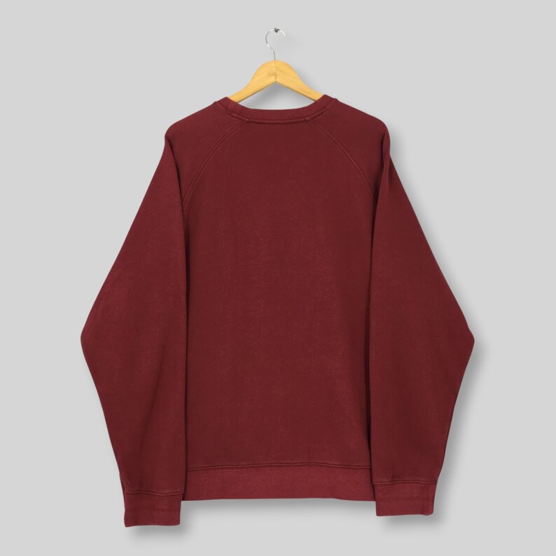 Może przedstawiać: Bordowa bluza z długimi rękawami, wisząca na drewnianym wieszaku. Bluza z okrągłym dekoltem jest w jednolitym kolorze i wydaje się być wykonana z miękkiego materiału. Odzież jest prezentowana na jasnoszarym tle.