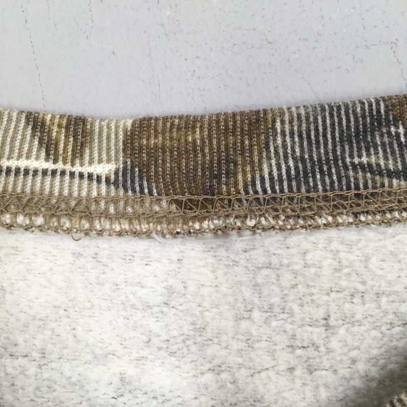 Op de afbeelding: Close-up van een camouflagepatroon stof, met tinten bruin, beige en grijs. De stof heeft een geribbelde kraag en een zachte, fleece binnenkant. Het ontwerp suggereert een kledingstuk, mogelijk een sweatshirt of iets dergelijks.