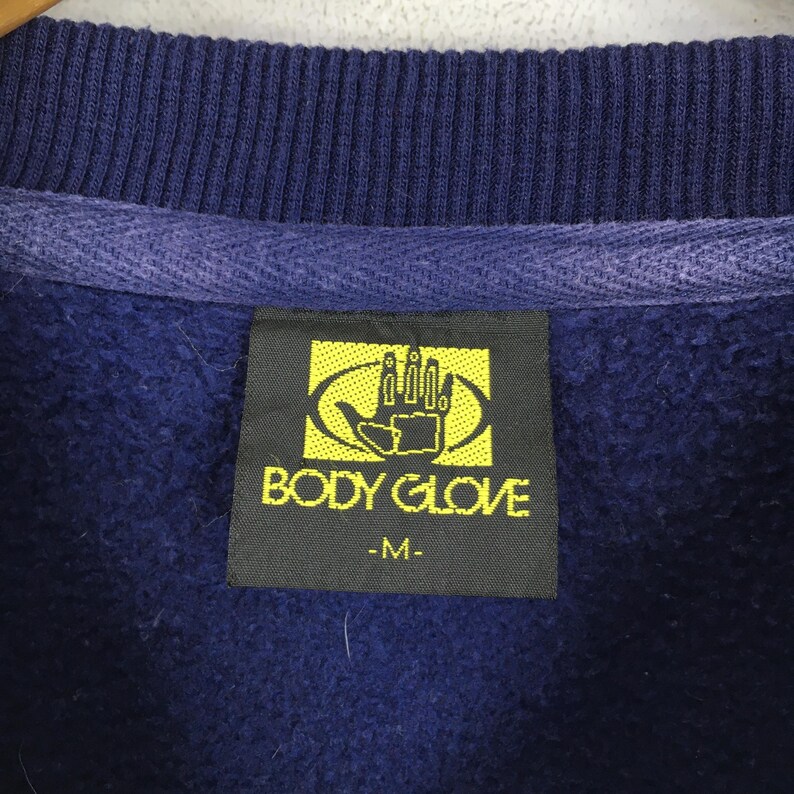 May include: A dark blue sweatshirt featuring a ribbed crew neckline and a black rectangular interior label. The label displays a yellow hand logo inside an oval, the text BODY GLOVE, and the size indicator M printed in yellow lettering.