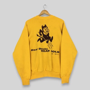 Puede incluir: Una sudadera amarilla con un gráfico de diablo marrón sosteniendo un tridente. El texto "Real Devils Wear Gold" está impreso debajo del gráfico. La sudadera está colgada de una percha de madera.