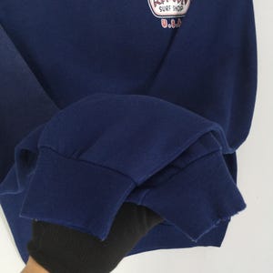 May include: A navy blue sweatshirt with a crew neck and ribbed cuffs. The upper left chest has a circular graphic with the text "Surf Shop U.S.A.". The sweatshirt is made of a soft, comfortable material.