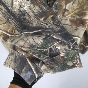 Op de afbeelding: Een camouflage-patroon sweatshirt in tinten bruin, groen en beige. De stof heeft een gedetailleerde print van bladeren en takken, met een geribde manchet zichtbaar aan de rand van de mouw. Het algehele ontwerp is een klassieke outdoor-kledingstijl.