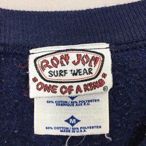 May include: A close-up of a navy blue sweatshirt collar with tags. The tags display "RON JON SURF WEAR ONE OF A KIND" and include fabric content and size details. The sweatshirt is made in the U.S.A.