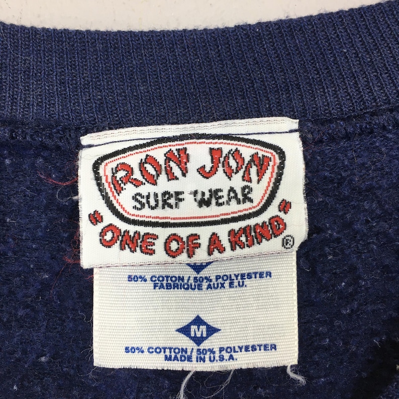 May include: A close-up of a navy blue sweatshirt collar with tags. The tags display "RON JON SURF WEAR ONE OF A KIND" and include fabric content and size details. The sweatshirt is made in the U.S.A.