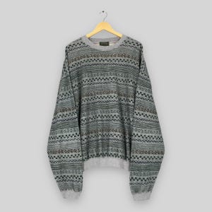 May include: A gray sweater with a geometric pattern in black, brown, and green. The sweater has a crew neck and long sleeves with ribbed cuffs. The garment is hanging on a wooden hanger against a light gray background.