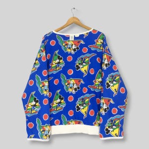 May include: A long-sleeve blue sweatshirt featuring an all-over print of Mickey Mouse windsurfing with colorful sails and red sun icons. The garment has a white crew neckline, white sleeve cuffs, and a white hemline, displayed on a wooden hanger.