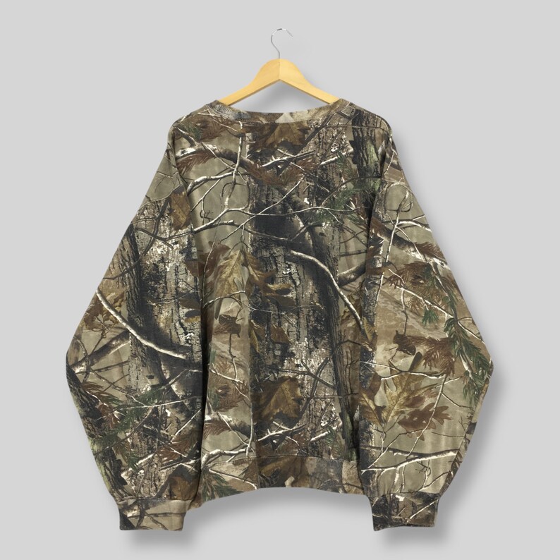 Op de afbeelding: Een camouflage sweatshirt met lange mouwen hangt aan een houten hanger. De sweatshirt heeft een realistisch bospatroon in bruin, groen en wit. Het heeft een ronde hals en geribde manchetten. Het kledingstuk is gemaakt van een zacht, comfortabel materiaal.
