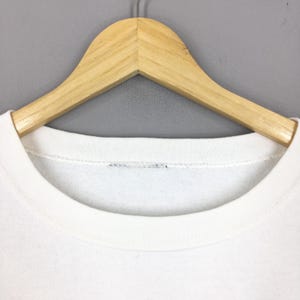 May include: A white crew neck sweatshirt hanging on a wooden hanger. The sweatshirt has a simple, clean design with a rounded neckline. The hanger is made of light-colored wood.
