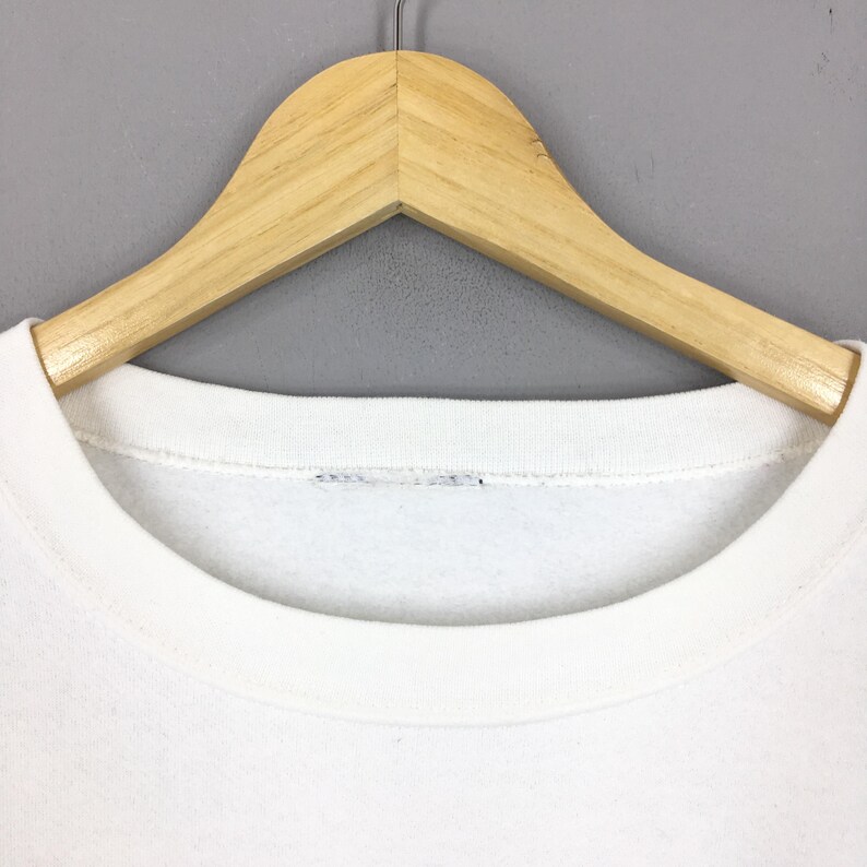 May include: A white crew neck sweatshirt hanging on a wooden hanger. The sweatshirt has a simple, clean design with a rounded neckline. The hanger is made of light-colored wood.