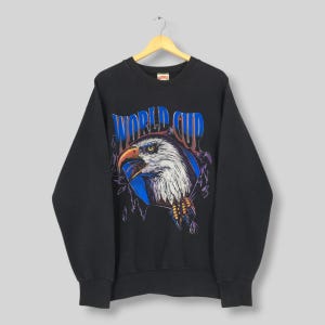 May include: Black sweatshirt featuring a graphic of a bald eagle's head breaking through a blue circle, with the words "WORLD CUP" in blue and white. The eagle has a yellow beak and talons. The sweatshirt is hanging on a wooden hanger.