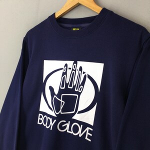 May include: A navy blue long-sleeve sweatshirt featuring a white graphic print on the chest. The graphic displays a stylised hand inside a white square and oval frame, with the text BODY GLOVE printed in white capital letters underneath the hand.