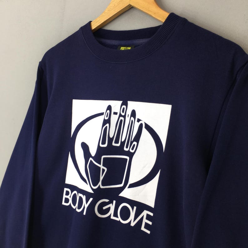 May include: A navy blue long-sleeve sweatshirt featuring a white graphic print on the chest. The graphic displays a stylised hand inside a white square and oval frame, with the text BODY GLOVE printed in white capital letters underneath the hand.