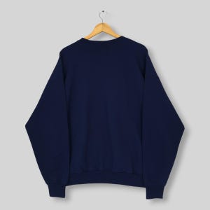 May include: A solid navy blue long-sleeve sweatshirt featuring a crew neckline, ribbed cuffs, and a ribbed hem. The garment is displayed on a wooden hanger, showcasing a plain, unadorned back view of the pullover with a consistent dark blue color.