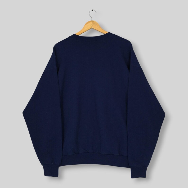 May include: A solid navy blue long-sleeve sweatshirt featuring a crew neckline, ribbed cuffs, and a ribbed hem. The garment is displayed on a wooden hanger, showcasing a plain, unadorned back view of the pullover with a consistent dark blue color.