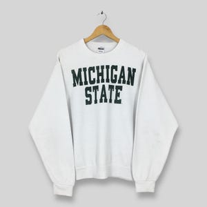 Puede incluir: Una sudadera blanca con la inscripción "MICHIGAN STATE" en letras de bloque verde oscuro. La prenda está colgada de una percha de madera. La sudadera tiene mangas largas y cuello redondo, sobre un fondo gris claro.