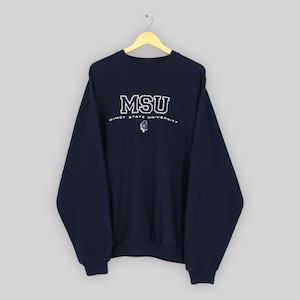 Puede incluir: Sudadera azul marino con las letras blancas "MSU" en un contorno punteado. Debajo, el texto "MINOT STATE UNIVERSITY" está impreso en blanco, con un pequeño gráfico. La sudadera está colgada de una percha de madera.