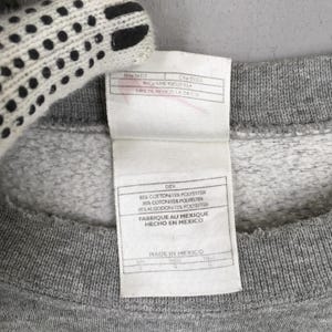 May include: Close-up of a gray sweatshirt with a white tag. The tag has text in English, French, and Spanish, including "85% COTTON/15% POLYESTER" and "MADE IN MEXICO". A white glove is partially visible in the upper left corner.