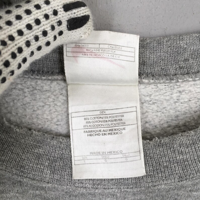 May include: Close-up of a gray sweatshirt with a white tag. The tag has text in English, French, and Spanish, including "85% COTTON/15% POLYESTER" and "MADE IN MEXICO". A white glove is partially visible in the upper left corner.