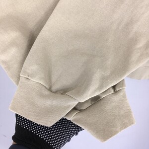 K&ouml;nnte beinhalten: Nahaufnahme eines beigefarbenen Langarm-Shirt-&Auml;rmels. Das B&uuml;ndchen ist gerippt und der Stoff scheint eine weiche Baumwollmischung zu sein. Der &Auml;rmel ist leicht zerknittert.