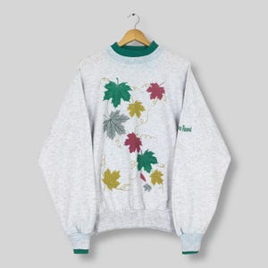 Op de afbeelding: Een lichtgrijs sweatshirt met een groene kraag en manchetten. Het sweatshirt heeft een geborduurd ontwerp van kleurrijke bladeren in groen, rood, geel en grijs. Het woord "Resort" staat op de mouw.