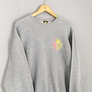 May include: A heather gray sweatshirt with a crew neck and long sleeves. The sweatshirt features a circular graphic on the chest with a hand design in yellow and pink. The garment is hanging on a wooden hanger.
