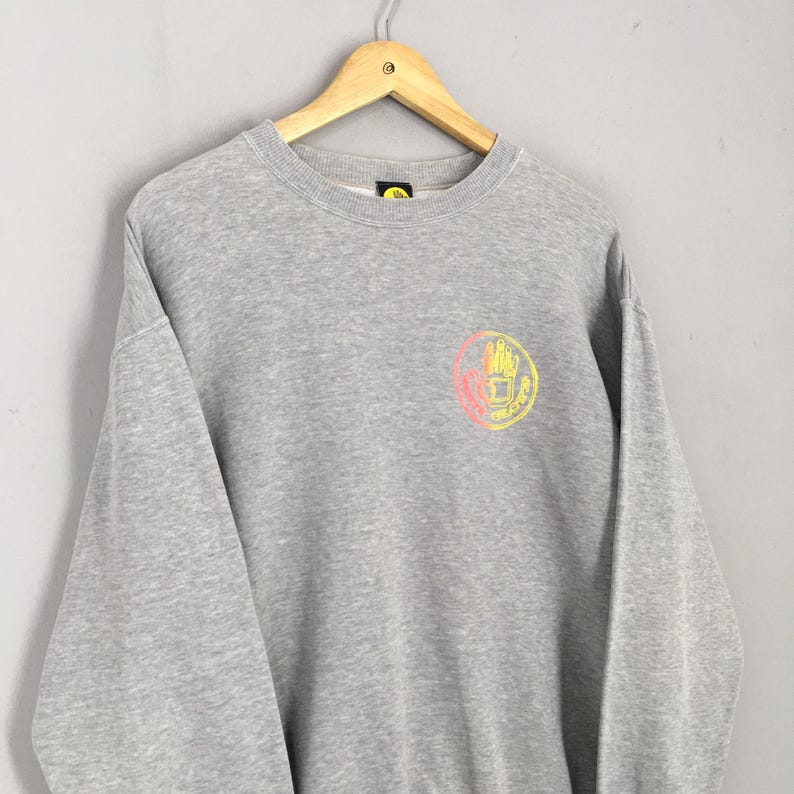 May include: A heather gray sweatshirt with a crew neck and long sleeves. The sweatshirt features a circular graphic on the chest with a hand design in yellow and pink. The garment is hanging on a wooden hanger.