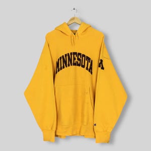 May include: Mustard yellow hoodie with the word "MINNESOTA" arched across the chest in dark brown lettering. The hoodie has a kangaroo pocket and a hood. The Champion logo is visible on the left sleeve.