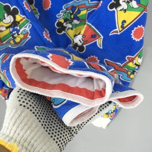 May include: A bright blue long-sleeve shirt featuring an all-over print of Mickey Mouse characters engaged in water sports. The design includes colorful sailboats, yellow triangles, and red starburst shapes. The cuffs are finished with white trim.