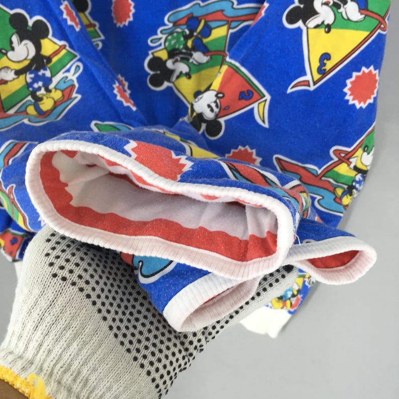 May include: A bright blue long-sleeve shirt featuring an all-over print of Mickey Mouse characters engaged in water sports. The design includes colorful sailboats, yellow triangles, and red starburst shapes. The cuffs are finished with white trim.