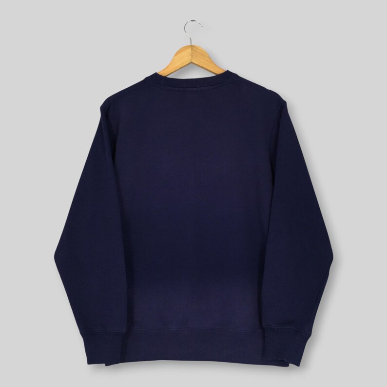 May include: A solid navy blue long-sleeve sweatshirt with a crew neckline, ribbed cuffs, and a ribbed hem. The garment is displayed on a wooden clothes hanger, showcasing a plain, unadorned back view of the dark blue fabric construction.