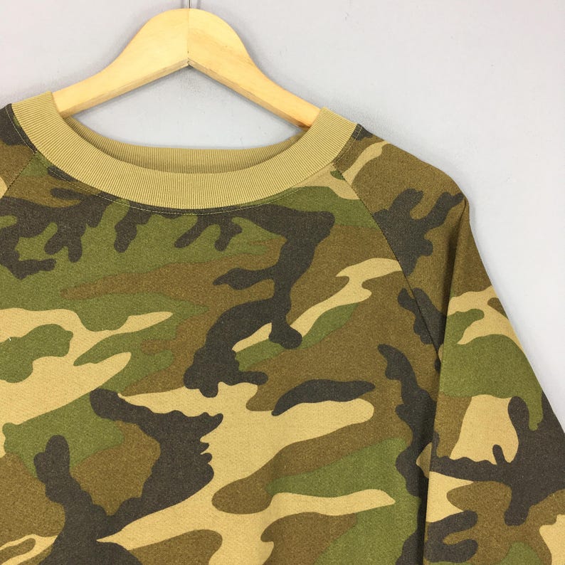 May include: A camouflage-patterned sweatshirt in shades of green, brown, and tan. The crew neck and raglan sleeves are visible. The sweatshirt is hanging on a wooden hanger against a neutral background.