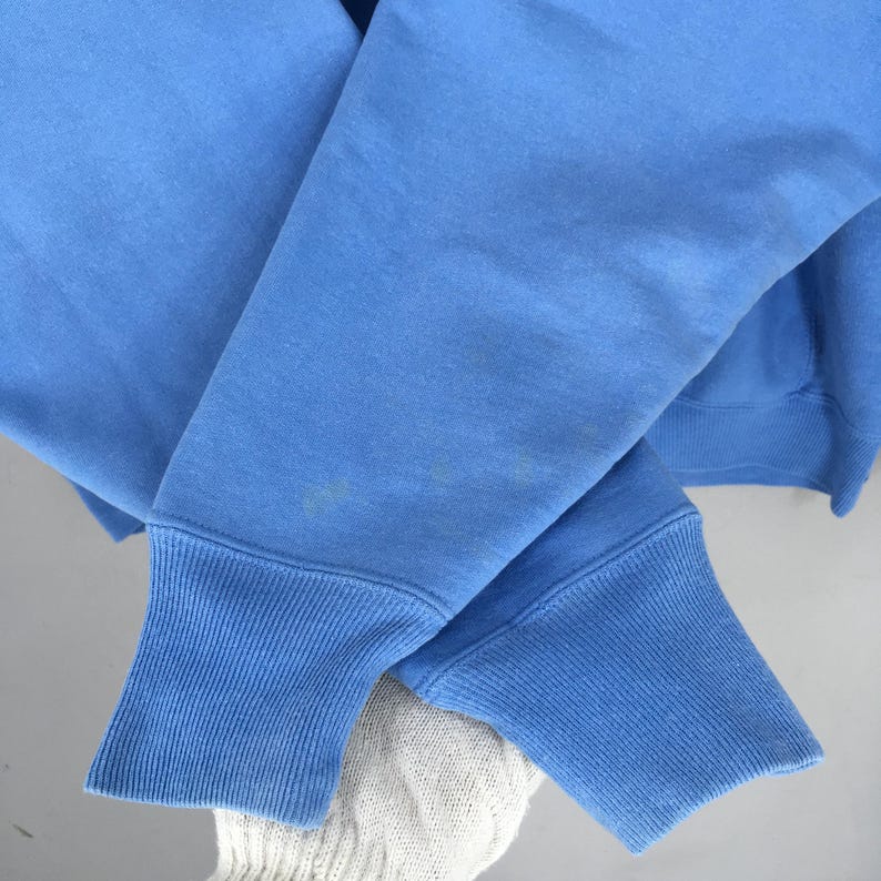 May include: Close-up of a light blue sweatshirt sleeve. The sleeve has a ribbed cuff and is made of a soft, comfortable material. The sweatshirt is a solid colour and appears to be well-worn.