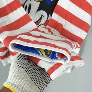 May include: A long-sleeve shirt featuring horizontal red and white stripes. The garment displays a graphic of Mickey Mouse on the chest. The interior of the sleeve cuff is lined with a blue fabric featuring a yellow triangle and the number 3.