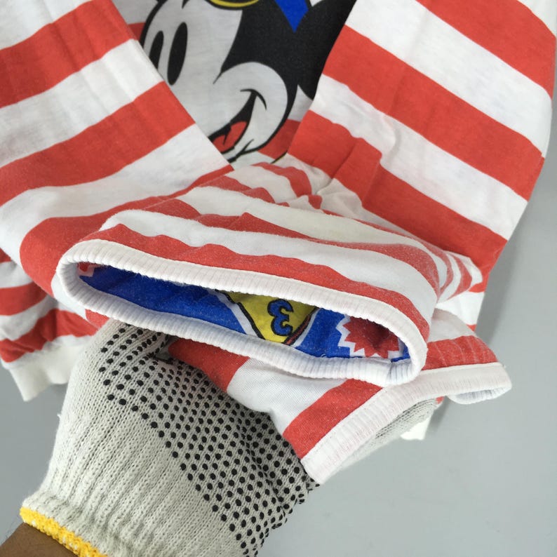 May include: A long-sleeve shirt featuring horizontal red and white stripes. The garment displays a graphic of Mickey Mouse on the chest. The interior of the sleeve cuff is lined with a blue fabric featuring a yellow triangle and the number 3.