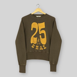 May include: Olive green sweatshirt with the number "25" and the word "SEAL" in a distressed yellow font. The sweatshirt has a crew neck and long sleeves. The garment is hanging from a wooden hanger.