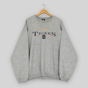 May include: A grey marl sweatshirt featuring the word "TIGERS" in navy blue and orange lettering. The sweatshirt has a crew neck and long sleeves. The Detroit Tigers logo is centered below the word "TIGERS". It is hanging on a wooden hanger.