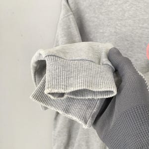 May include: Close-up of a gray sweatshirt sleeve with ribbed cuffs. The sleeve is folded over, revealing the inside fabric. The cuff is a lighter shade of gray than the rest of the sleeve.