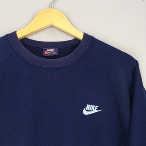 May include: A navy blue Nike sweatshirt with a crew neck and long sleeves. The Nike logo is embroidered in white on the chest. The sweatshirt is hanging on a wooden hanger.