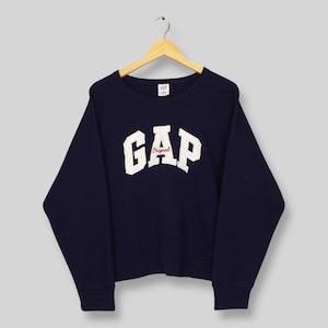 May include: A navy blue GAP sweatshirt with a crew neck and long sleeves. The white "GAP" logo is prominently displayed on the front, with "Original" in red script underneath. The sweatshirt is hanging on a wooden hanger.