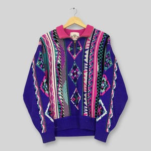 May include: A colourful vintage sweater in purple, pink, teal, and white. The sweater has a zip-up collar and a geometric pattern with diamond and stripe designs. The sweater is hanging on a wooden hanger.