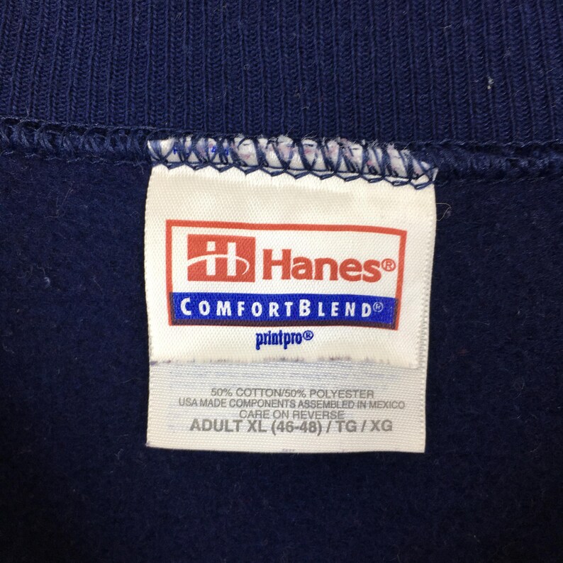 May include: A navy blue garment featuring a white Hanes ComfortBlend tag. The label displays the Hanes logo, 50% cotton and 50% polyester fabric composition, USA made components assembled in Mexico, and the size Adult XL (46-48).