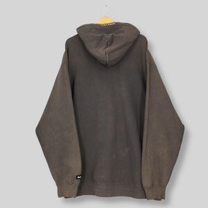 Puede incluir: Una sudadera con capucha marr&oacute;n con capucha con cord&oacute;n, mangas largas y un bolsillo de canguro. La sudadera con capucha tiene un aspecto descolorido y un peque&ntilde;o logotipo en la manga izquierda. La capucha tiene texto blanco.