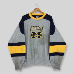 Könnte beinhalten: Ein graues Sweatshirt mit Rundhalsausschnitt, marineblauen und gelben Ärmeln im Color-Blocking-Design und einem gelben Kragen. Auf der Brust befindet sich der gestickte Schriftzug MICHIGAN über einem großen, marineblau-gelben M-Blockbuchstaben.
