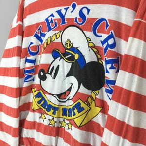 May include: A long-sleeve shirt featuring horizontal red and white stripes. The graphic print displays Mickey Mouse wearing a sailor hat with an anchor icon, surrounded by the blue text Mickey's Crew and a yellow banner reading First Mate.