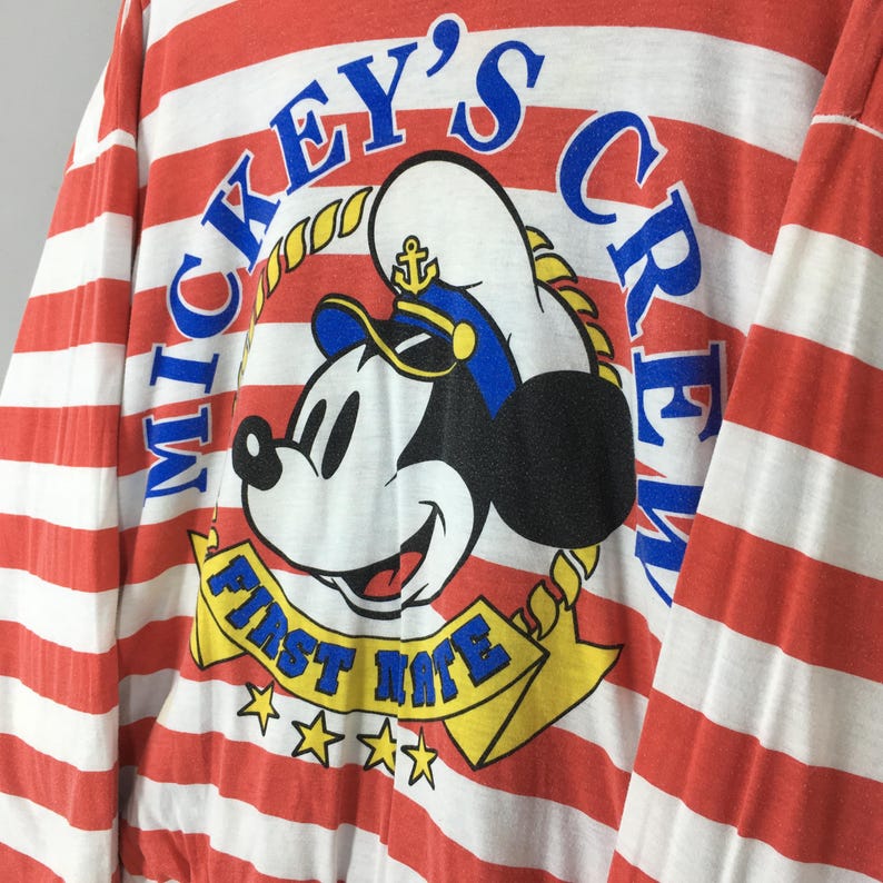 May include: A long-sleeve shirt featuring horizontal red and white stripes. The graphic print displays Mickey Mouse wearing a sailor hat with an anchor icon, surrounded by the blue text Mickey's Crew and a yellow banner reading First Mate.