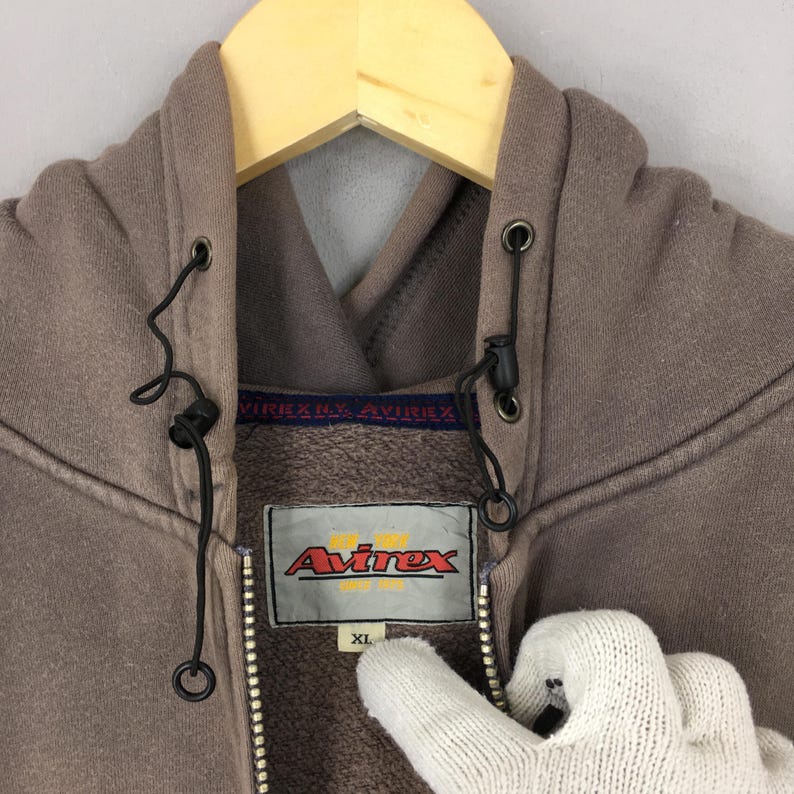 Puede incluir: Una sudadera con capucha marr&oacute;n con cremallera y capucha con cord&oacute;n, con el logotipo "Avirex" en una etiqueta rectangular. La sudadera tiene cierre de cremallera y una etiqueta XL. La prenda est&aacute; colgada en una percha de madera.