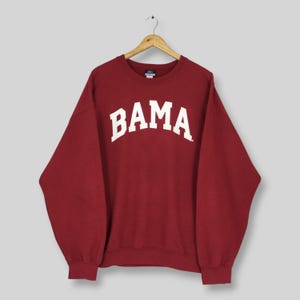 May include: A maroon long-sleeve crewneck sweatshirt featuring the word BAMA printed in large white capital letters across the chest in an arched font. The garment is displayed on a wooden hanger against a plain light gray background.