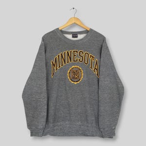 May include: A charcoal grey crewneck sweatshirt featuring the word MINNESOTA in arched yellow and black lettering across the chest. Below the text is a circular University of Minnesota emblem in yellow and black. The garment has long sleeves.