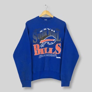 以下が含まれることがあります： 「Super Bowl」の文字が赤色で、その下に「Bills」と書かれたロイヤルブルーのスウェットシャツ。バッファローのグラフィックと「1992 AFC Champions」の文字が特徴です。木製のハンガーに吊るされています。