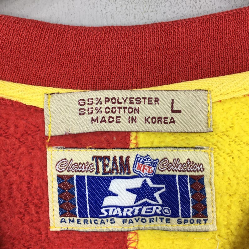 May include: Close-up of a red, yellow, and orange sweatshirt tag. The tag reads "65% Polyester, 35% Cotton, Made in Korea" and features the Starter and NFL logos. The text "America's Favorite Sport" is also visible.